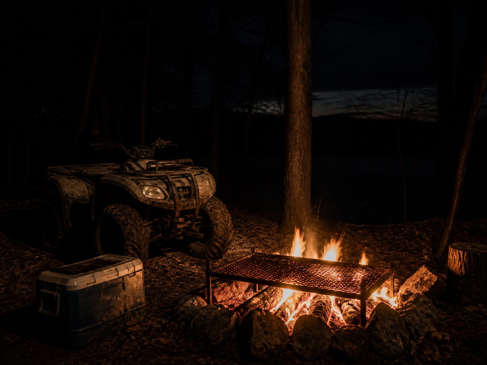 Muddy ATV parked beside a campfire with a grill grate over coals